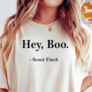May include: A white t-shirt with the text "Hey, Boo. - Scout Finch" printed on the front. The shirt is being worn by a person wearing black biker shorts.