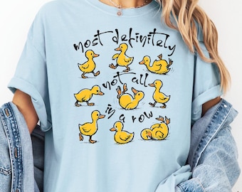 May include: Light blue t-shirt with a graphic of yellow cartoon ducks in various poses. The text "Most definitely not all in a row" is written above the ducks in black script. The shirt is part of the Chambray collection.