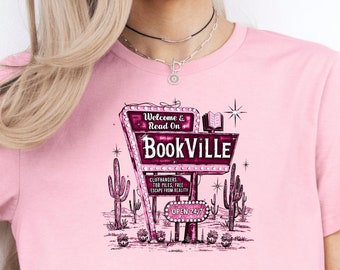 May include: Pink t-shirt with a graphic of a retro sign that says "Welcome & Read On BookVILLE" with cacti and stars. The sign has text that says "Cliffhangers, TBR Piles, Free Escape From Reality, Open 24/7".