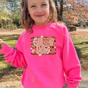 May include: A bright pink sweatshirt with the words "Game Day" in a decorative design. The graphic includes footballs, flowers, and other sports-themed elements. The sweatshirt is a vibrant color and appears to be made of a soft material.