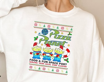 May include: White sweatshirt featuring a graphic of the Pizza Planet logo and three green aliens wearing Santa hats. The text reads "FOOD & FUN SPACE PORT SERVING YOUR LOCAL STAR CLUSTER". The design includes Christmas-themed elements.