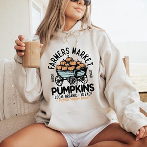 May include: A cream-colored hoodie with a graphic that reads "Farmers Market Pumpkins." The graphic features a wagon filled with pumpkins. The hoodie is worn by a person sitting on a wooden bench.