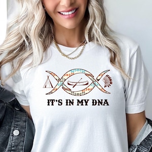 May include: White t-shirt with a graphic of a canoe, a teepee, and a headdress inside a double helix shape. The text "It's in my DNA" is below the graphic.