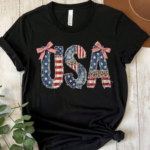 May include: A black short-sleeved t-shirt featuring the text USA in a distressed, patchwork-style font. The letters are decorated with red, white, and blue flag patterns, leopard print accents, and small red and white striped bows on the U and A.