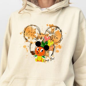 May include: Cream-colored hoodie featuring a graphic of the Orange Bird character with a Mickey Mouse ear design. The design includes orange and yellow accents, with the text "Orange Bird" below the character. The hoodie has a front pocket and a drawstring hood.