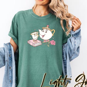 May include: Light green t-shirt featuring a cartoon teapot and teacup design with a pink rose and a book. The shirt has the words "Light Green" in a script font. The shirt is a relaxed fit.