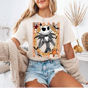 May include: A cream-colored t-shirt featuring a graphic print of Jack Skellington from The Nightmare Before Christmas inside an oval frame with orange stripes, spiderwebs, bats, and stars. Six additional design options are displayed as thumbnails.