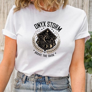 Puede incluir: Una camiseta blanca con un dise&ntilde;o gr&aacute;fico en negro y dorado que presenta un drag&oacute;n, lunas y estrellas. El gr&aacute;fico tiene el texto "ONYX STORM" y "BRAVE THE DARK".