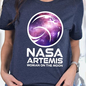 May include: Navy blue t-shirt featuring a circular graphic with a galaxy design and the text "NASA ARTEMIS WOMAN ON THE MOON" in white. The shirt has short sleeves and a crew neck.