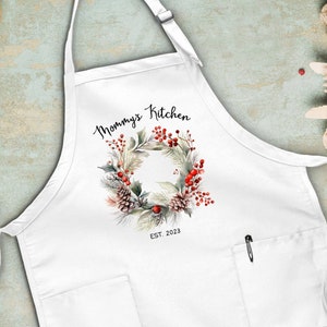 May include: White kitchen apron with a red and green wreath design and the text "Mommy's Kitchen Est. 2023".