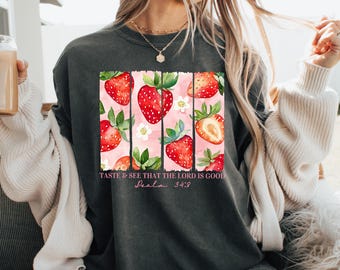 May include: Dark gray t-shirt featuring a strawberry design. The design includes red strawberries, white flowers, and the text "Taste & See That The Lord Is Good" from Psalm 34:8. The shirt is a comfortable, casual style.