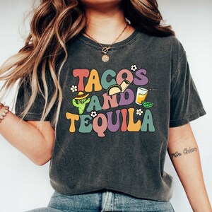 Puede incluir: Una camiseta gris oscuro con un gr&aacute;fico colorido que dice "Tacos and Tequila" con un cactus, tacos y una copa de margarita.
