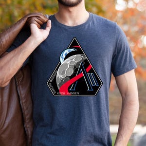 May include: A dark gray t-shirt featuring a triangular patch with a space-themed design. The patch includes a depiction of the moon, Earth, and the names "WISEMAN", "GLOVER", "KOCH", and "HANSEN". A brown leather jacket is draped over the person's shoulder.