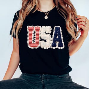 May include: Black t-shirt with the word "USA" in red and white striped and navy blue letters. The letters are outlined with a white lace trim. The shirt is worn with dark wash jeans and white sneakers.