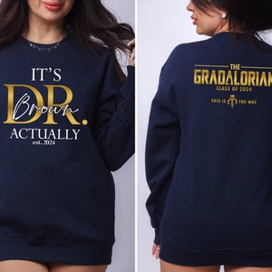 May include: Navy blue sweatshirt with gold text that reads "IT'S DR. Brown ACTUALLY est. 2024" on the front and "THE GRADALORIAN CLASS OF 2024 THIS IS THE WAY" on the back.