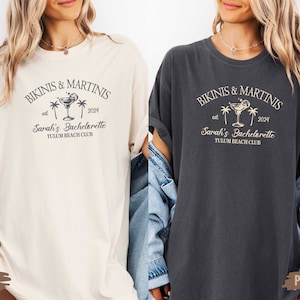 May include: Two women wearing oversized t-shirts with the text "Bikinis & Martinis est 2024 Sarah's Bachelorette Tulum Beach Club" and a martini glass with palm trees graphic. The shirt on the left is ivory and the shirt on the right is pepper.