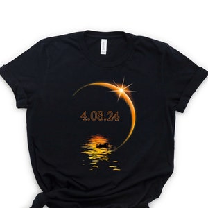 May include: Black t-shirt with an orange graphic of a solar eclipse with the date 4.08.24 printed on it.
