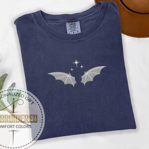 May include: Navy blue Comfort Colors t-shirt with an embroidered bat and star design. The shirt features a crew neck and short sleeves. The design includes a bat with outstretched wings and three stars.
