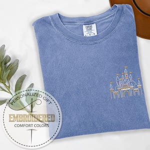 May include: A light blue t-shirt with an embroidered outline of a castle in pink, yellow, and white. The text "Personalized Gift" and "Embroidered Comfort Colors" is printed on a white circle.