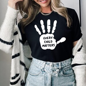May include: Black t-shirt with a white handprint graphic and the text "EVERY CHILD MATTERS". The shirt is made of a soft material and is suitable for everyday wear. The shirt is paired with blue denim jeans.