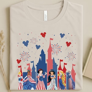 May include: Beige t-shirt with a patriotic design, featuring a red and blue castle silhouette, fireworks, and Mickey Mouse balloons. The shirt displays Disney princesses in red, white, and blue dresses. The word "CANVAS" is printed on the shirt.