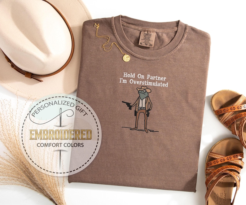 May include: A brown Comfort Colors t-shirt with the embroidered phrase "Hold On Partner I'm Overstimulated" and a cartoon frog wearing a cowboy hat and holding a gun. A gold necklace and a beige hat are next to the shirt. Brown sandals are also in the image.