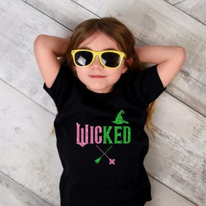 May include: A black t-shirt with the word "Wicked" in pink and green glitter, along with a witch hat and crossed broomsticks. The person in the image is wearing yellow sunglasses.