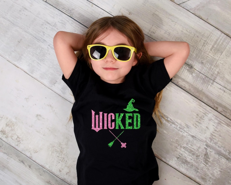 May include: A black t-shirt with the word "Wicked" in pink and green glitter, along with a witch hat and crossed broomsticks. The person in the image is wearing yellow sunglasses.