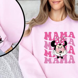 Puede incluir: Sudadera rosa con un gr&aacute;fico de Minnie Mouse con gafas de sol y la palabra "MAMA" repetida en una fuente retro. Las mangas tienen un peque&ntilde;o gr&aacute;fico bordado de Minnie Mouse con los nombres "Oliver" y "Sarah" en los pu&ntilde;os.