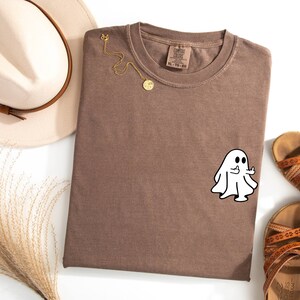 May include: A brown t-shirt with a white ghost giving the middle finger. The ghost is outlined in black. The t-shirt is folded and laid flat on a white surface.