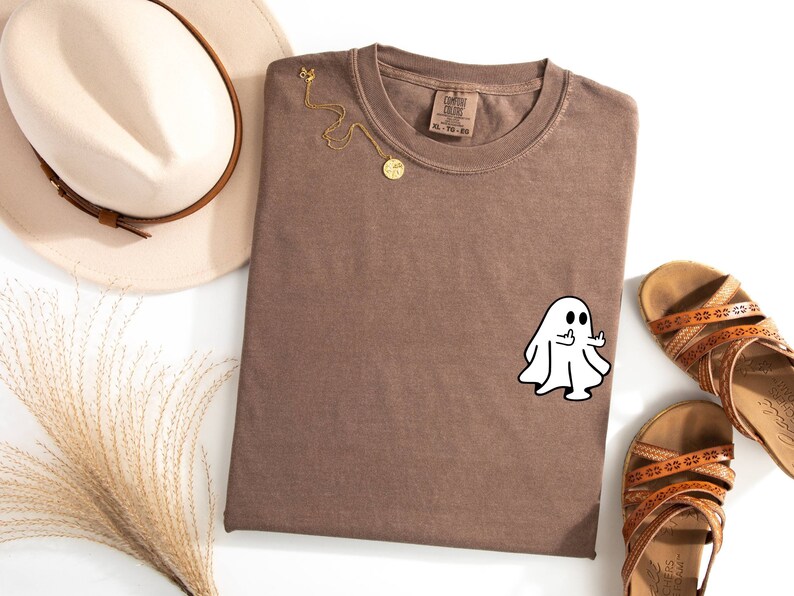 May include: A brown t-shirt with a white ghost giving the middle finger. The ghost is outlined in black. The t-shirt is folded and laid flat on a white surface.