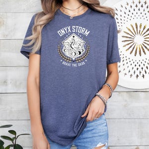 Puede incluir: Una camiseta azul marino con un gr&aacute;fico de un drag&oacute;n en un c&iacute;rculo con el texto "ONYX STORM" y "BRAVE THE DARK" impreso en la parte delantera.