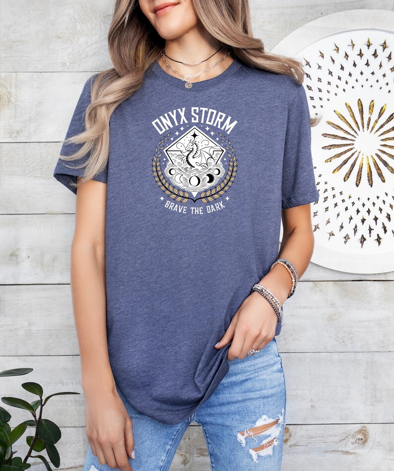 Puede incluir: Una camiseta azul marino con un gr&aacute;fico de un drag&oacute;n en un c&iacute;rculo con el texto "ONYX STORM" y "BRAVE THE DARK" impreso en la parte delantera.