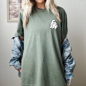 May include: A green t-shirt with a white ghost graphic giving a thumbs up. The ghost has two black eyes.