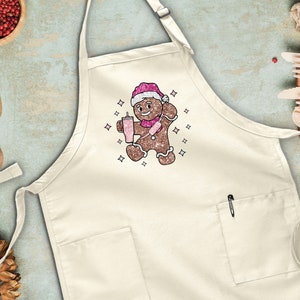Peut inclure: Un tablier blanc avec un motif d'homme en pain d'&eacute;pices. L'homme en pain d'&eacute;pices porte un chapeau de P&egrave;re No&euml;l et tient une tasse de chocolat chaud. Le tablier a une poche sur le devant et une boucle pour l'accrocher.