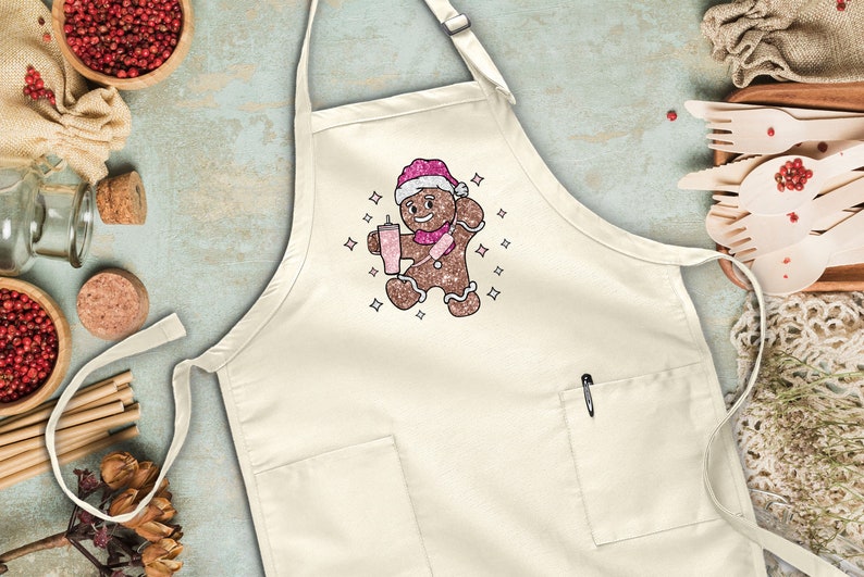 Peut inclure: Un tablier blanc avec un motif d'homme en pain d'&eacute;pices. L'homme en pain d'&eacute;pices porte un chapeau de P&egrave;re No&euml;l et tient une tasse de chocolat chaud. Le tablier a une poche sur le devant et une boucle pour l'accrocher.