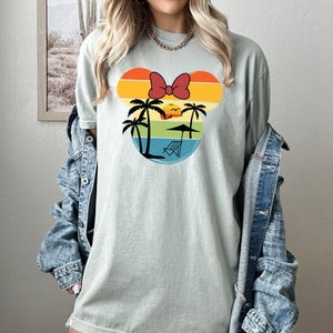 May include: A light green t-shirt with a retro-style graphic of a Mickey Mouse head silhouette. The graphic features a sunset with palm trees, a beach umbrella, and a beach chair. The Mickey Mouse ears are made of colorful stripes and a red bow.