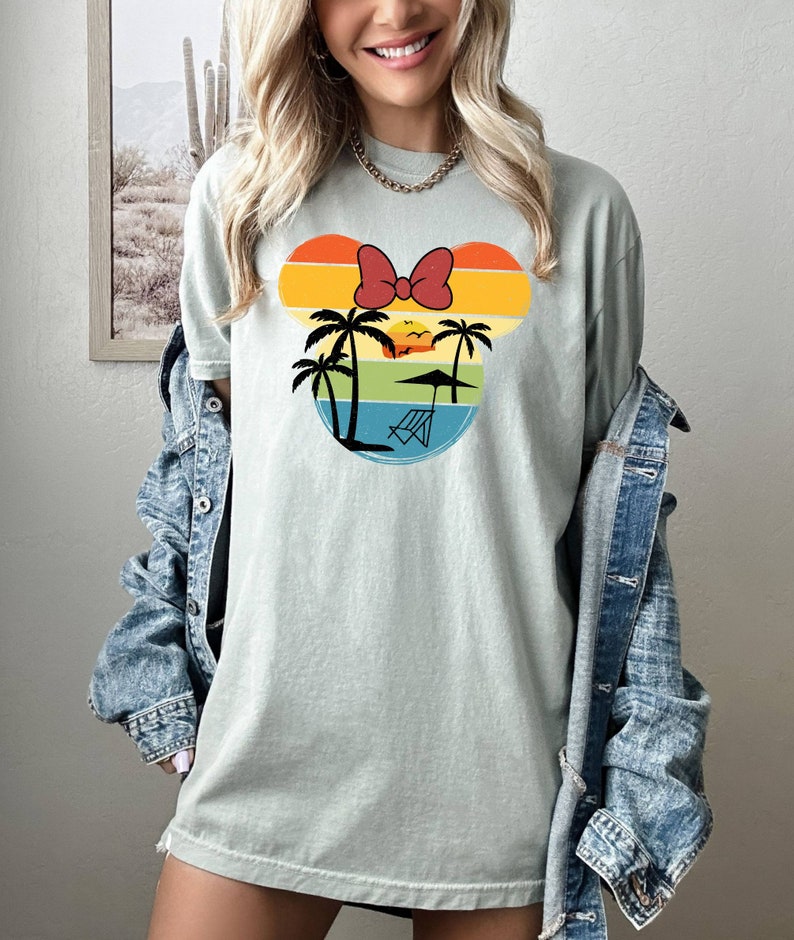 May include: A light green t-shirt with a retro-style graphic of a Mickey Mouse head silhouette. The graphic features a sunset with palm trees, a beach umbrella, and a beach chair. The Mickey Mouse ears are made of colorful stripes and a red bow.