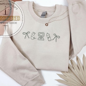 Peut inclure: Un sweat-shirt beige avec des dessins brod&eacute;s d'un n&oelig;ud, d'une image d'&eacute;chographie, d'un ordinateur, d'un flacon pulv&eacute;risateur et d'un n&oelig;ud. Un collier dor&eacute; avec un pendentif est sur le sweat-shirt. Le texte "EMBROIDERED SWEATSHIRT & HOODIE" est dans un cercle.