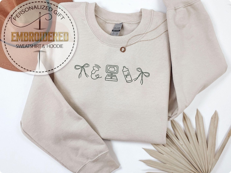 Peut inclure: Un sweat-shirt beige avec des dessins brod&eacute;s d'un n&oelig;ud, d'une image d'&eacute;chographie, d'un ordinateur, d'un flacon pulv&eacute;risateur et d'un n&oelig;ud. Un collier dor&eacute; avec un pendentif est sur le sweat-shirt. Le texte "EMBROIDERED SWEATSHIRT & HOODIE" est dans un cercle.