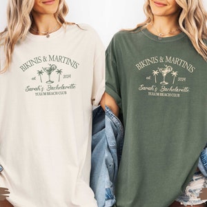May include: Two oversized t-shirts with a vintage-inspired design. The shirts are in ivory and moss green colors. The design features a martini glass with a lime wedge, palm trees, and the text "Bikinis & Martinis est 2024 Sarah's Bachelorette Tulum Beach Club".
