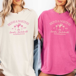 May include: Two women wearing t-shirts with the text "Bikinis & Martinis est 2024 Sarah's Bachelorette Tulum Beach Club" in pink. The t-shirt on the left is ivory and the t-shirt on the right is pink.