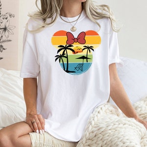 May include: White t-shirt with a retro-style graphic of a Mickey Mouse head silhouette. The graphic features a sunset, palm trees, a beach umbrella, and a lounge chair.