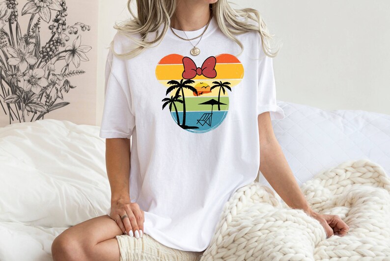 May include: White t-shirt with a retro-style graphic of a Mickey Mouse head silhouette. The graphic features a sunset, palm trees, a beach umbrella, and a lounge chair.