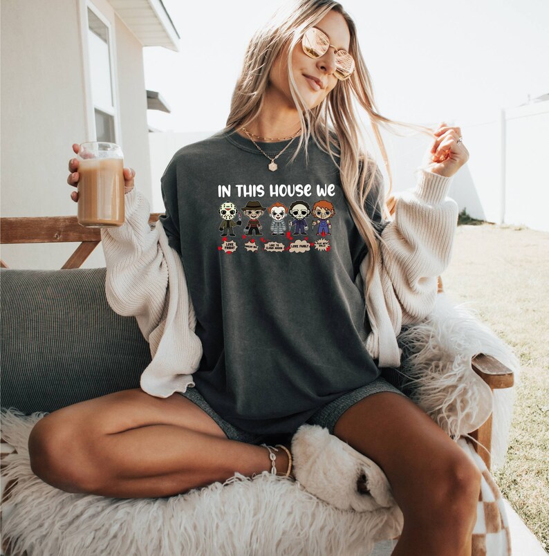 May include: A dark gray sweatshirt with a graphic print of horror movie characters. The text "In this house we" is printed above the characters. Each character has a speech bubble with a different phrase. The phrases are "Scream", "Think", "Love Family", "Live Freaky", and "Die Happy".