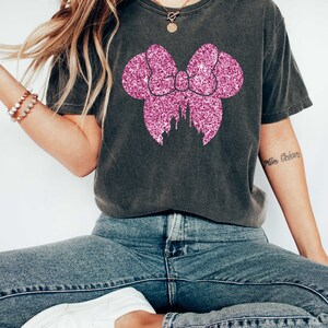May include: A dark gray t-shirt with a pink glitter bow and castle silhouette. The bow is in the shape of Mickey Mouse ears.