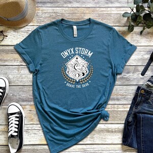 Puede incluir: Una camiseta azul turquesa con un dise&ntilde;o gr&aacute;fico blanco que presenta un drag&oacute;n, lunas y estrellas. El texto "Onyx Storm" y "Brave the Dark" est&aacute; impreso en la camiseta.
