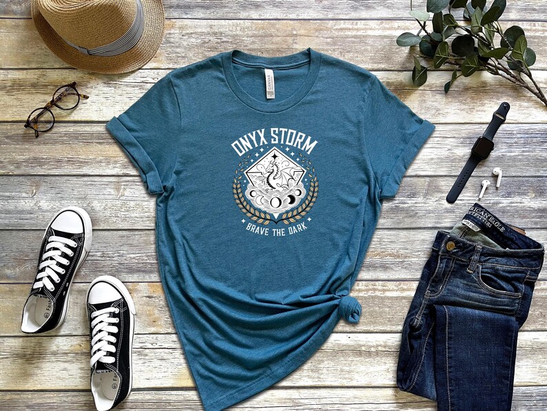 Puede incluir: Una camiseta azul turquesa con un dise&ntilde;o gr&aacute;fico blanco que presenta un drag&oacute;n, lunas y estrellas. El texto "Onyx Storm" y "Brave the Dark" est&aacute; impreso en la camiseta.