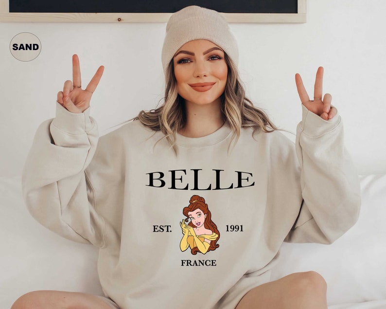 Peut inclure: Sweat-shirt couleur sable avec le mot "BELLE" en noir, et une illustration de Belle de La Belle et la B&ecirc;te. Le sweat-shirt comprend &eacute;galement "EST. 1991 FRANCE" en texte noir. Le mod&egrave;le porte un bonnet beige.