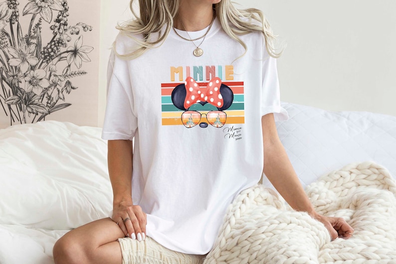 Op de afbeelding: Wit T-shirt met een retro-stijl grafische afbeelding van Minnie Mouse met een zonnebril en een rode strik. De afbeelding bevat de tekst "Minnie" en een regenboogstrepenpatroon.